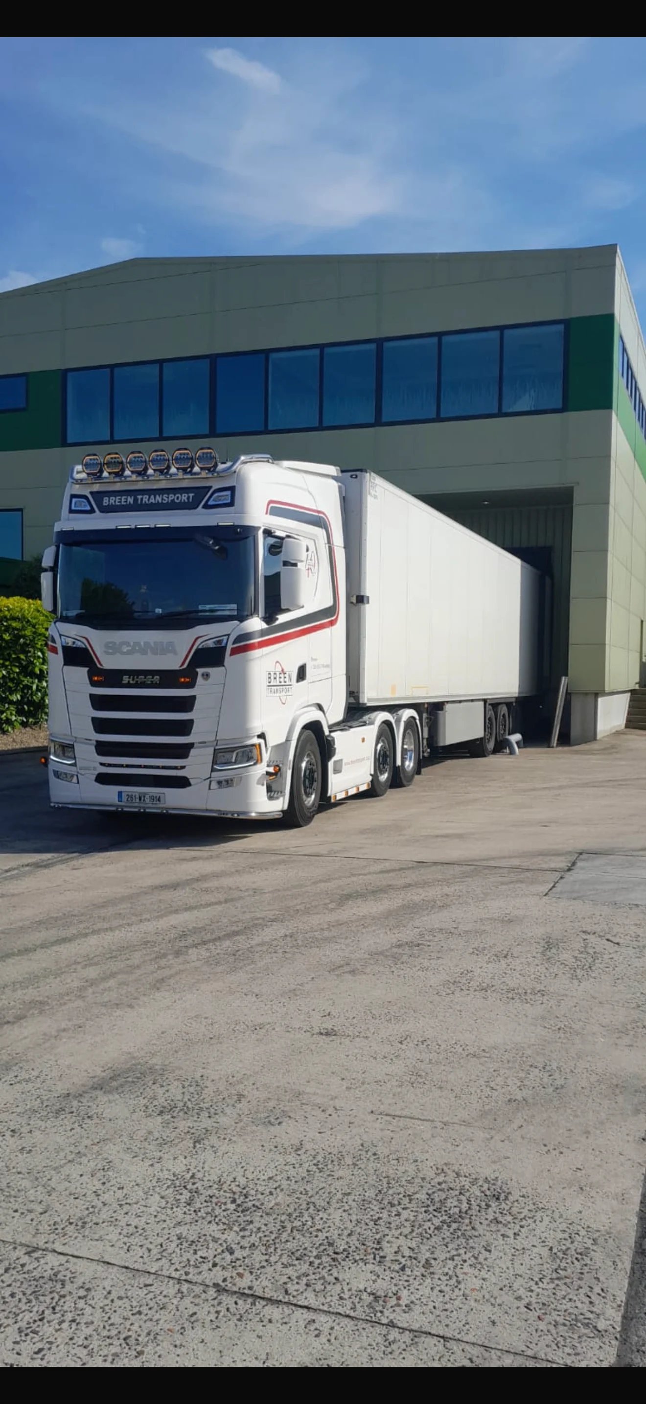 White Scania semi-truck with refrigerated trailer parked at a modern industrial building with blue windows