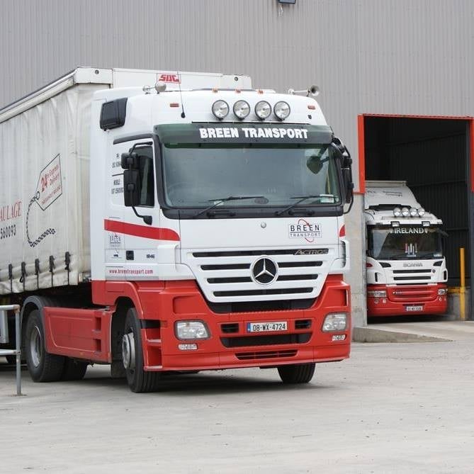 Red and white Mercedes articulated truck with BREHN TRANSPORT branding parked at a loading dock with another truck visible